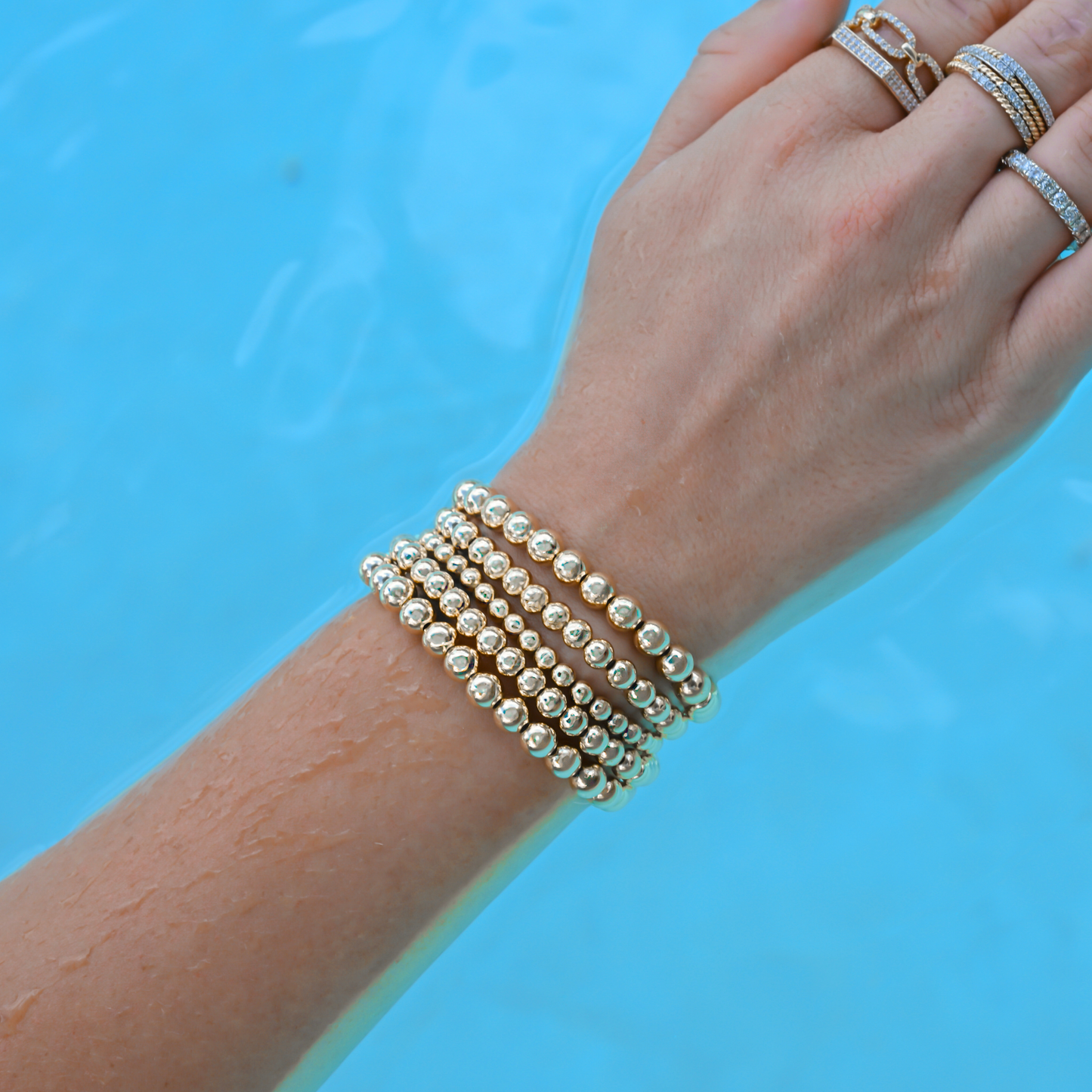 Hand wearing multiple gold-filled beaded bracelets from Gold Club by Shannon Hudson stacked on a woman’s wrist in a pool.