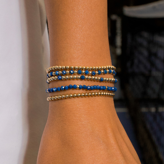 Gold-filled beaded bracelets from Gold Club by Shannon Hudson stacked on a woman’s wrist on a tennis and pickleball court.