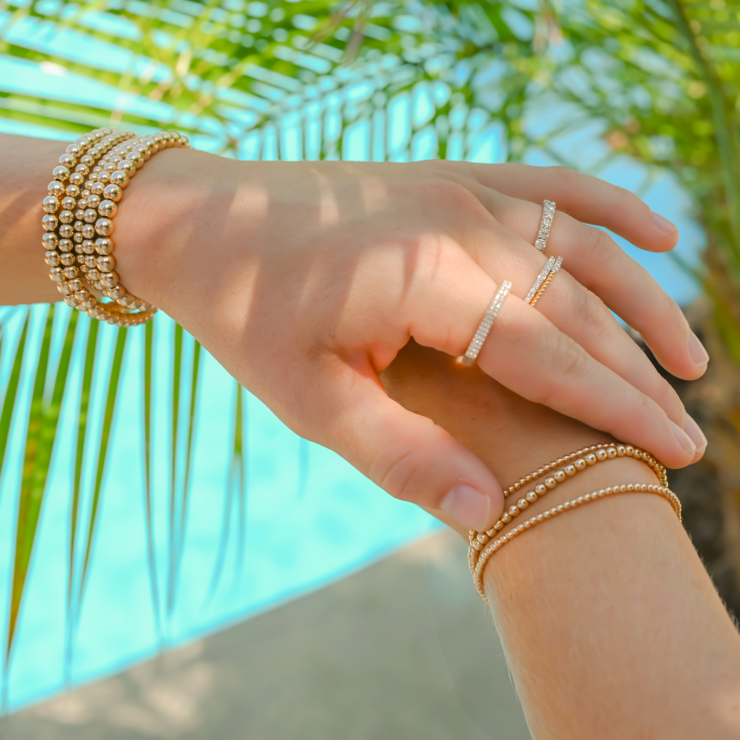 Hand wearing stacks of Gold Club by Shannon Hudson Gold LustraFlex™ Bracelets and rings with a blurred palm leaf and water background