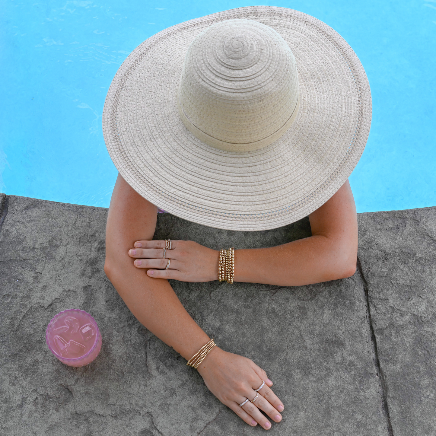 Person wearing a wide-brimmed hat and gold filled jewelry by a poolside with a pink cocktail on a stone ledge.