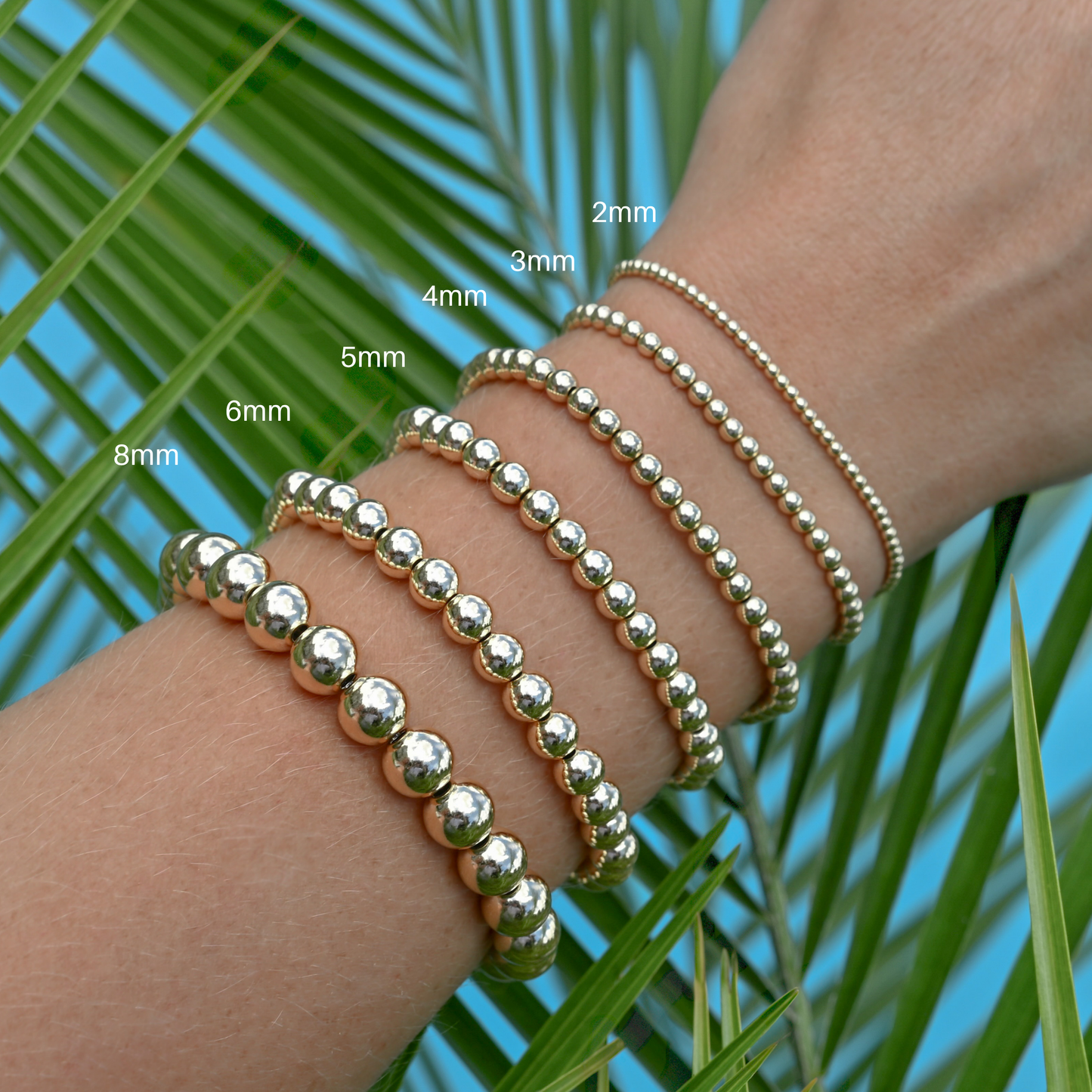Gold filled LustraFlex™ bracelets on a wrist with size labels against a palm leaf background