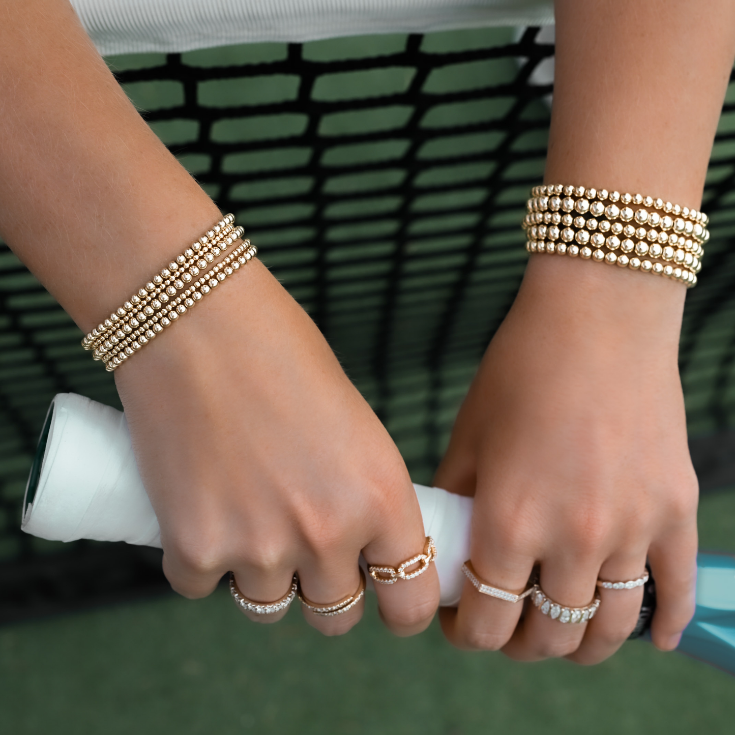 Close-up of hands wearing Gold LustraFlex™ bracelets and solid gold and diamond rings on a tennis background