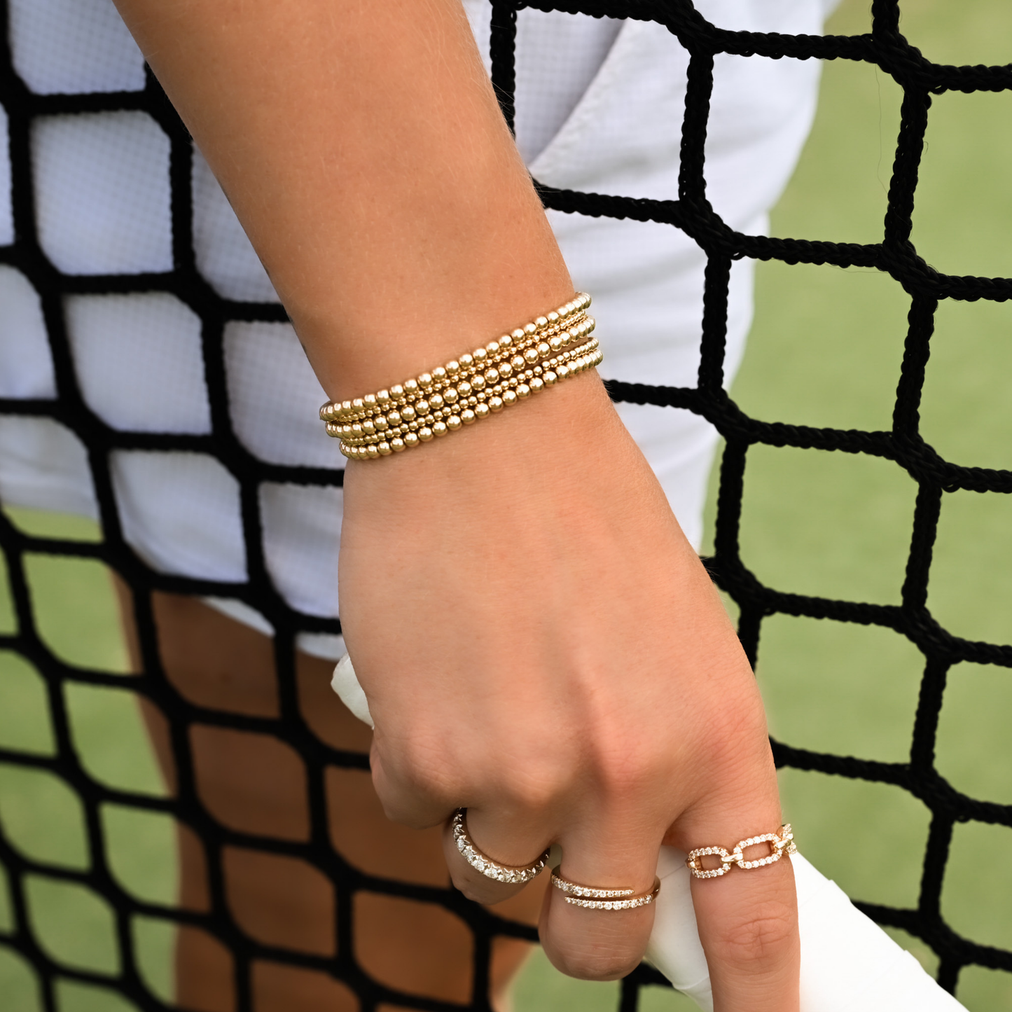Hand wearing gold bracelets and rings with a tennis background