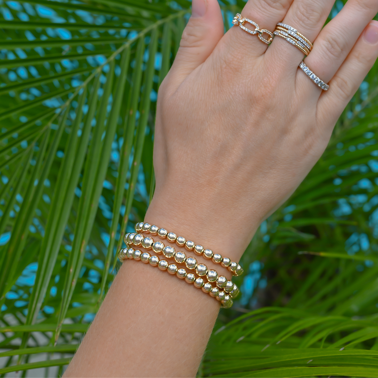 LustraFlex™ gold-filled beaded bracelet and rings by Gold Club on a palm leaf background.