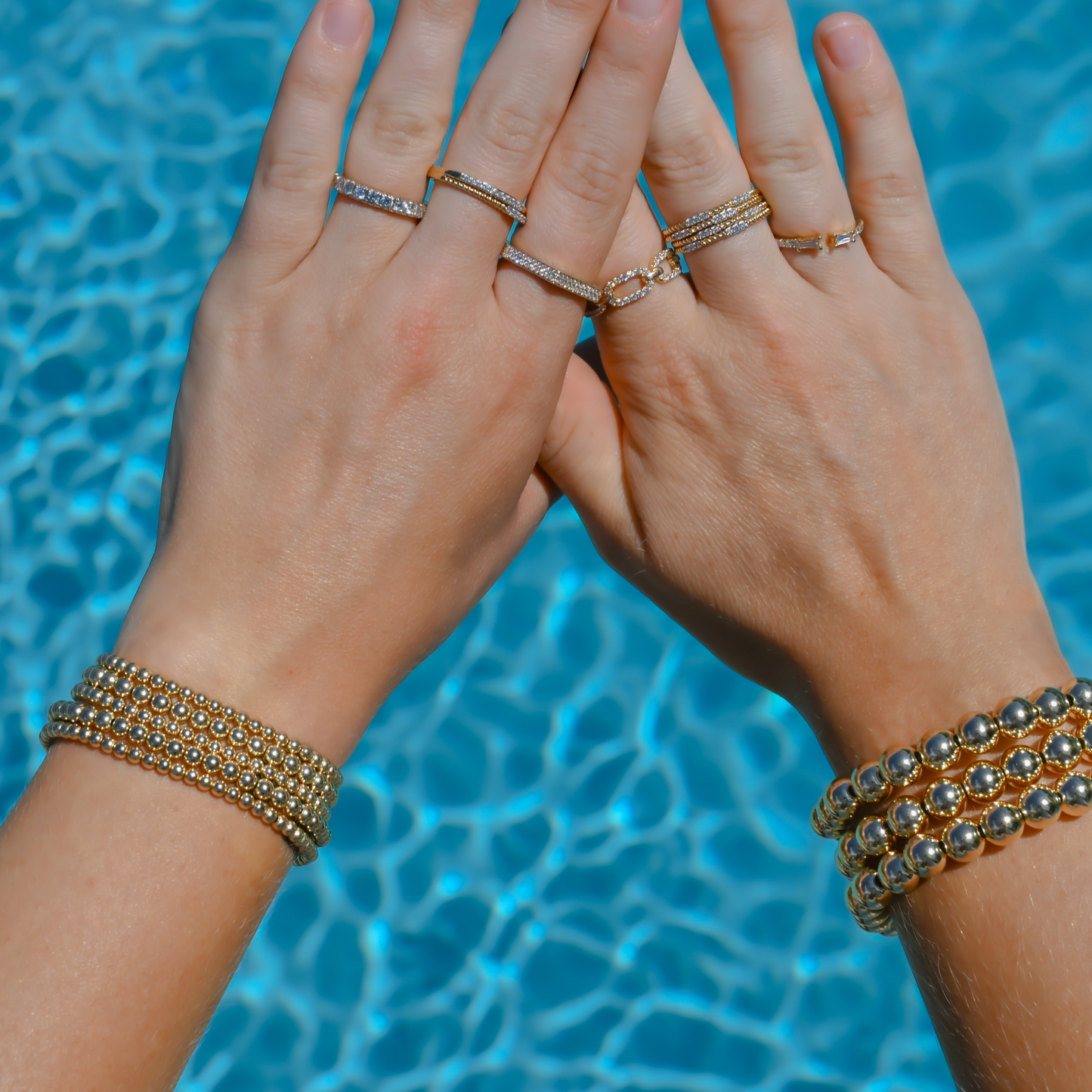 Two hands with gold filled bracelets and diamond rings against a blue water background