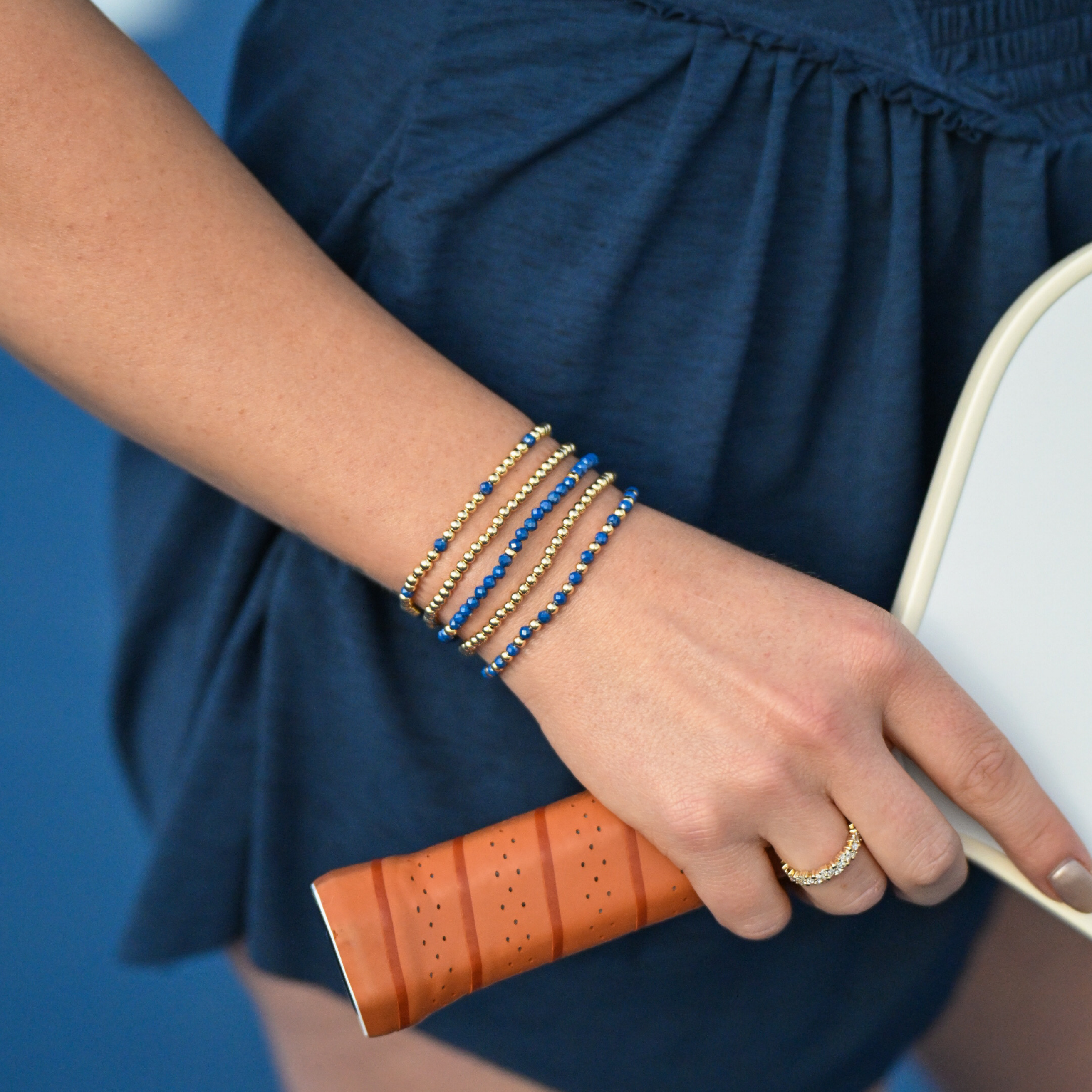 Close-up of the Gold Club Blue Zircon Signature Stack with layered blue zircon and gold beaded bracelets styled on a tennis court against a tennis racket backdrop.
