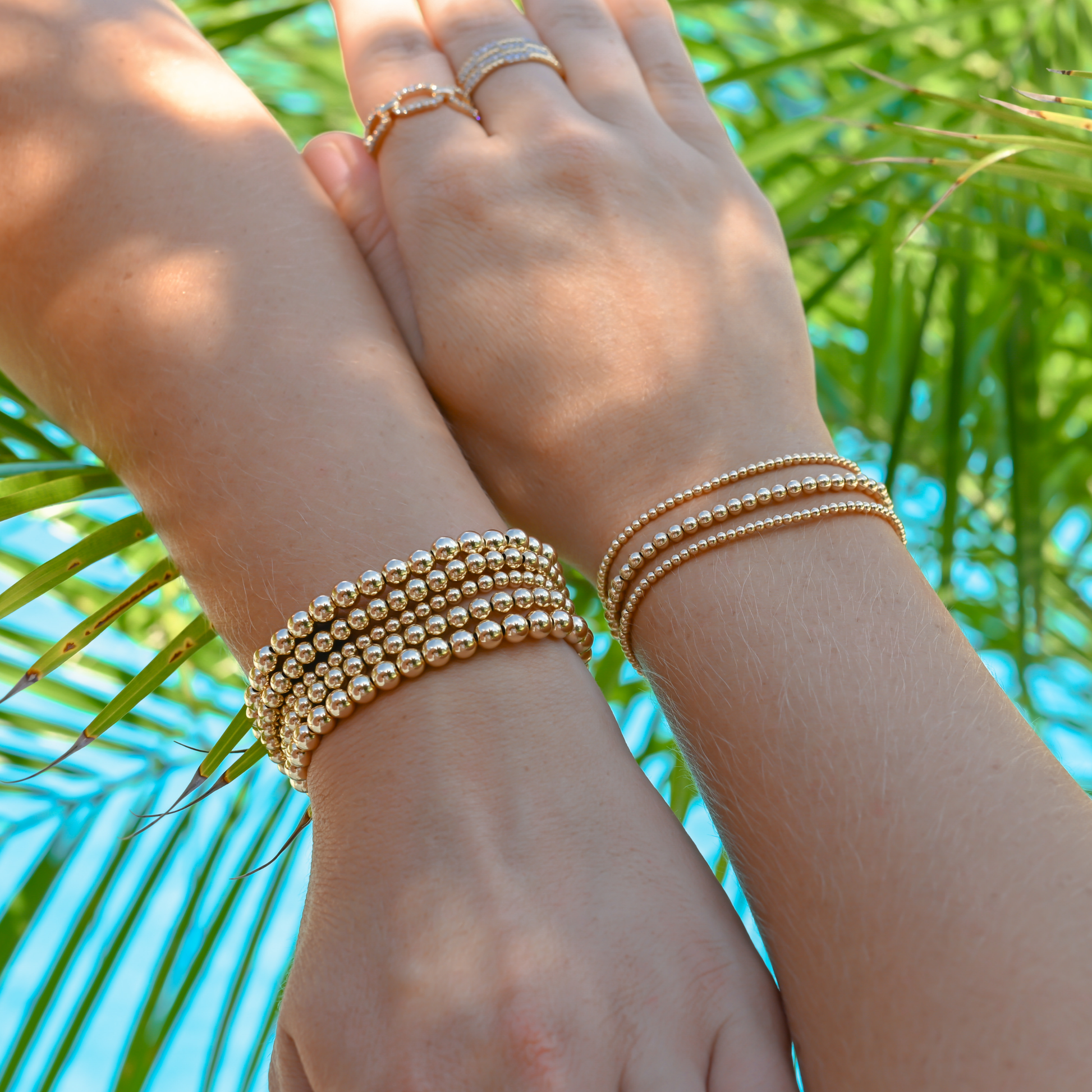Two hands wearing LustraFlex™ gold-filled beaded bracelet by Gold Club and rings against a palm leaf background