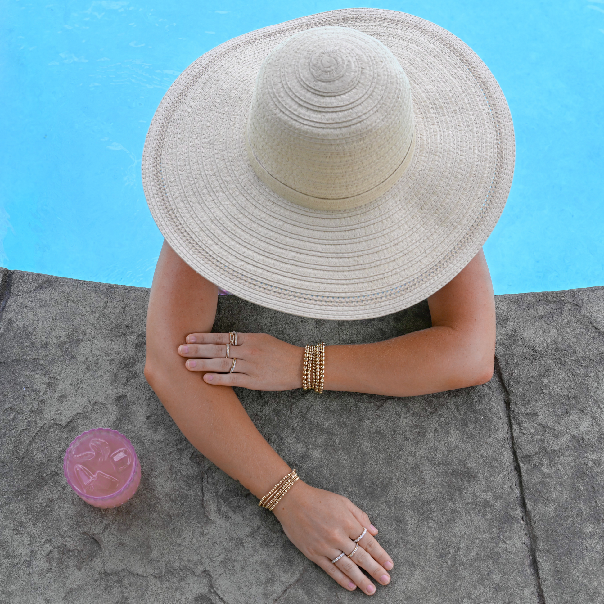 Person wearing a wide-brimmed hat and gold filled jewelry by a poolside with a pink cocktail on a stone ledge.