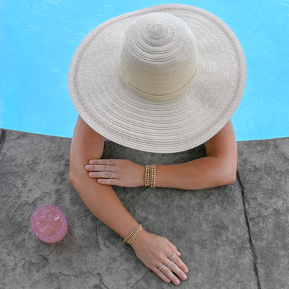 Person wearing a wide-brimmed hat and gold filled jewelry by a poolside with a pink cocktail on a stone ledge.