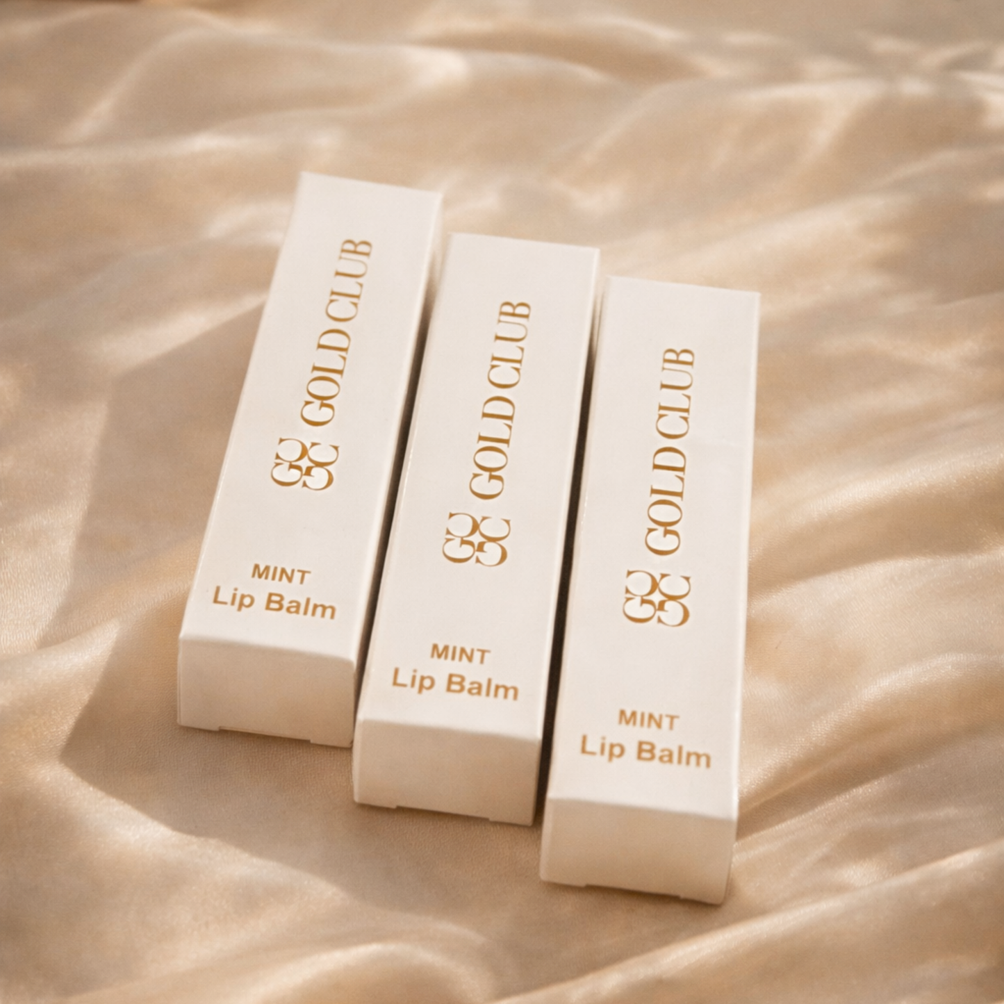Three Gold Club Mint Lip Balm containers on a beige fabric background