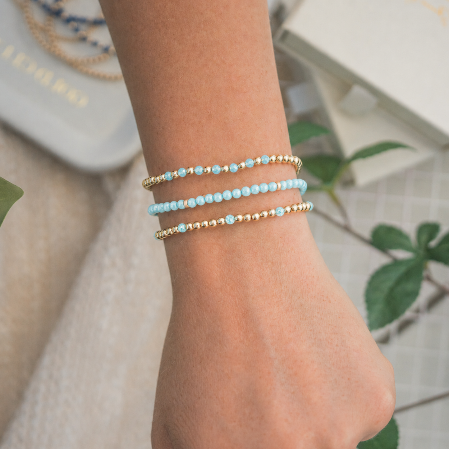 Three delicate beaded wrap bracelets on a person's wrist, featuring light blue zircon beads and small gold spacer beads, shown against a softly blurred background with green leaves. Sweat proof and tarnish resistant.