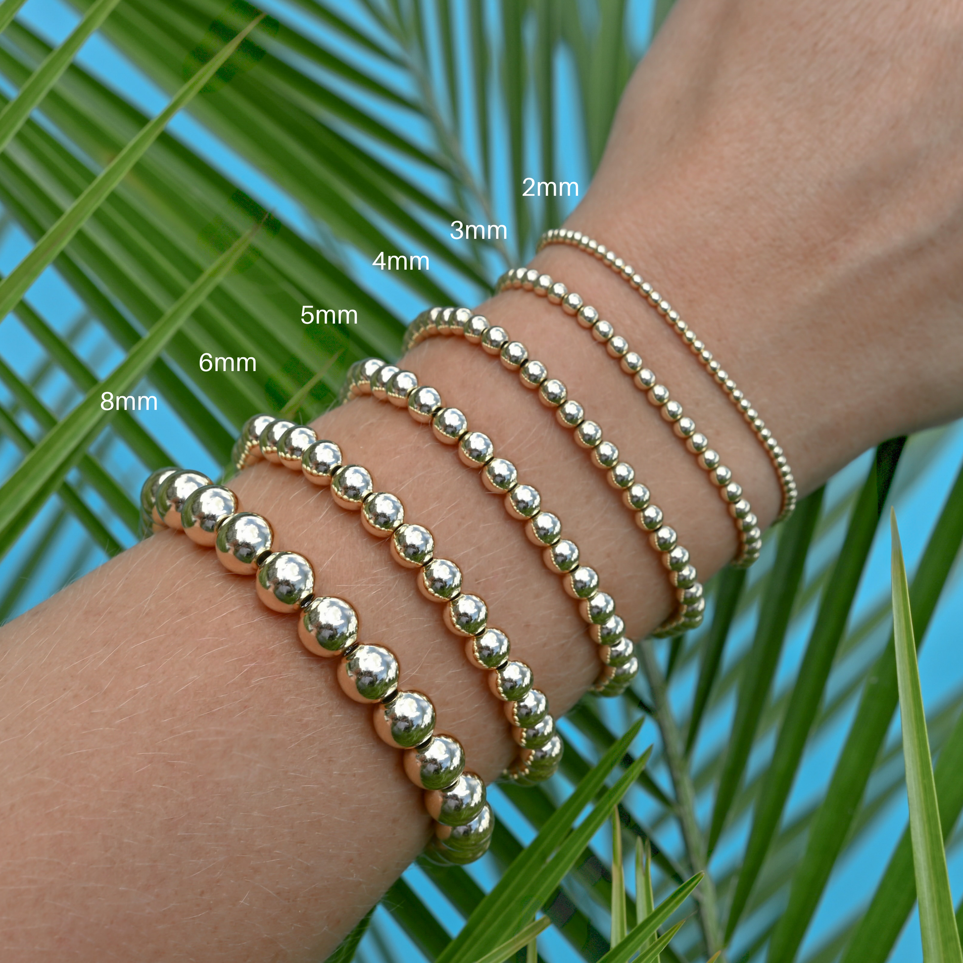 Gold filled LustraFlex™ bracelets on a wrist with size labels against a palm leaf background