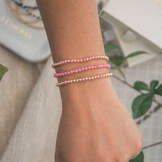 Three beaded stretch bracelets on a person's wrist, featuring pink and gold beads, shown in a styled lifestyle setting with a neutral background and green leaves. Swat proof and tarnish resistant.