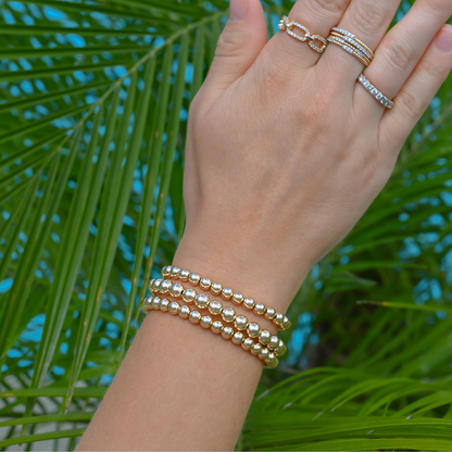 LustraFlex™ gold-filled beaded bracelet and rings by Gold Club on a palm leaf background.