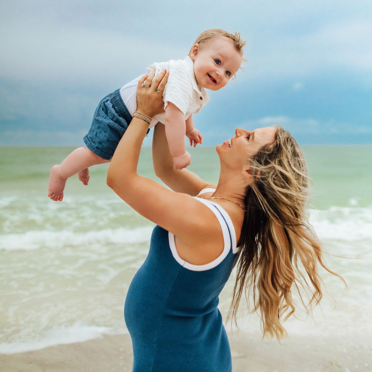 A mom holding a baby up in the air during a beach photoshoot for Gold Club by Shannon Hudson