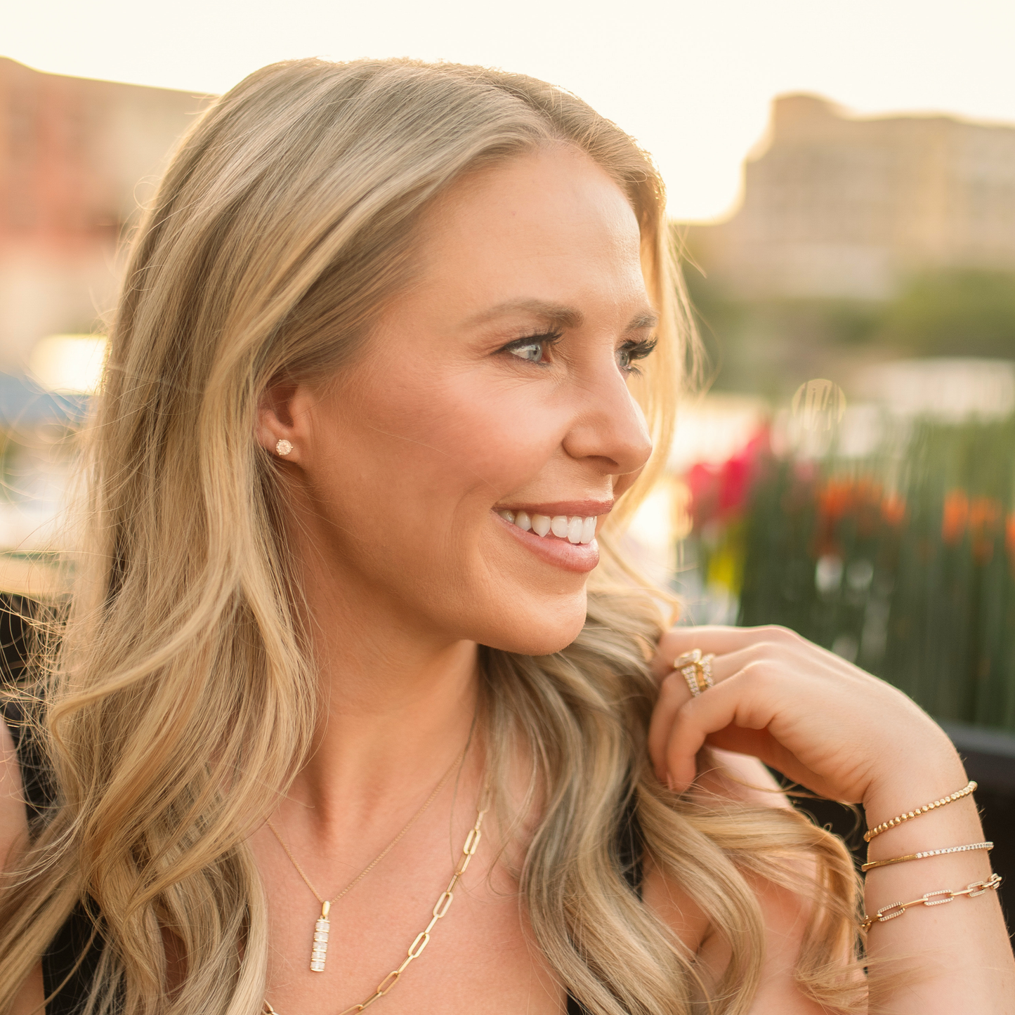 A smiling blonde woman in profile wearing delicate gold jewelry, including a small stud earring, a thin chain necklace with a rectangular pendant, and stacked bracelets, set against a softly blurred outdoor background. Jewelry from Gold Club Jewelry House on Shannon Hudson