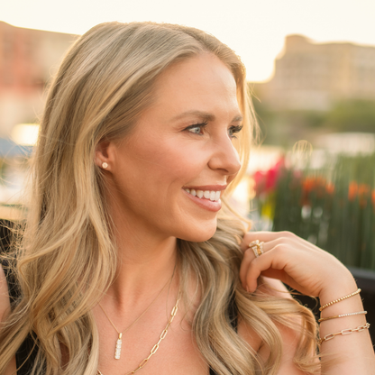 A smiling blonde woman in profile wearing delicate gold jewelry, including a small stud earring, a thin chain necklace with a rectangular pendant, and stacked bracelets, set against a softly blurred outdoor background. Jewelry from Gold Club Jewelry House on Shannon Hudson