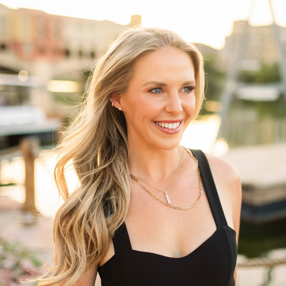 A smiling blonde woman wearing a black sleeveless top and layered gold necklaces, photographed outdoors with a softly blurred waterfront or dock background at warm golden hour. 1 carat diamond studs from Gold Club Jewelry House on Shannon Hudson at a cafe