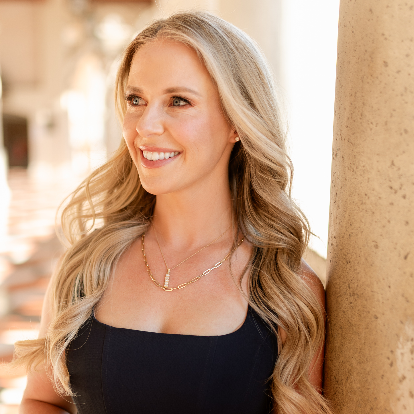 A smiling blonde woman with wavy hair leans against a stone column, wearing a black sleeveless top and layered gold necklaces in a warm, sunlit architectural setting. Jewelry from Gold Club Jewelry House on Shannon Hudson