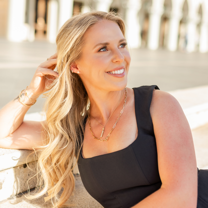 A smiling blonde woman in a black sleeveless top and gold necklaces, leaning against a stone wall in a sunlit outdoor setting. Jewelry from Gold Club Jewelry House on Shannon Hudson.