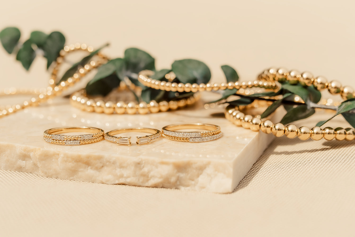 Gold Club by Shannon Hudson jewelry including rings and bracelets on a beige surface with green leaves.