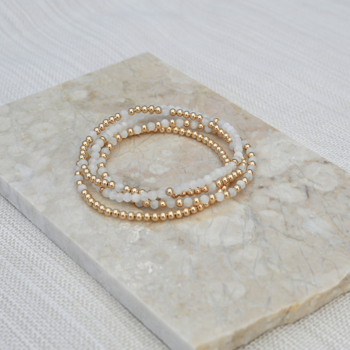 Stacking white moonstone gemstone bracelets from Gold Club highlight on a beige tile